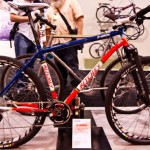 A colorful mountain bike displayed at a cycling exhibition. The bike features a blue and red frame with the brand "Ritchey" prominently marked. It includes thick tires suitable for off-road use, a front suspension fork, and is positioned on a black display pedestal. In the background, blurred figures and additional bicycles can be seen, indicating a busy event.