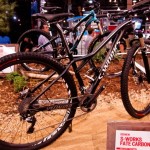 A black and blue mountain bike displayed on a sandy and rocky surface, showcasing its sleek design and features, including the brand name "Specialized" on the frame. In the background, blurred figures and trees are visible, highlighting an exhibition setting. A sign with "S-Works Fate Carbon 29" is positioned near the bike.