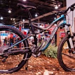 A mountain bike on display, featuring a sleek blue and black frame, thick tires for rugged terrain, and a visible gear system. The bike is positioned on a natural wood and gravel surface with a blurred background of an indoor exhibition space.