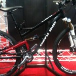 A black and red mountain bike displayed on a stand, featuring a prominent frame design, equipped with front suspension forks and a rear shock. The bike has a sleek appearance, showcasing its tires and details such as the gearing system. The background includes a grid structure, suggesting an exhibit or showcase setting.