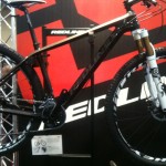 A modern mountain bike from Redline, displayed prominently on a stand. The bike features a sleek black frame, high-performance tires, and a fork with suspension. The background includes a large Redline logo, enhancing the bike's branding. An informational placard is visible beneath the bike.