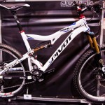 Alt text: A white Pivot mountain bike displayed at a bike expo, featuring a sleek design with red and blue accents, large tires, and a prominent brand logo. The backdrop is dark, emphasizing the bike's details.
