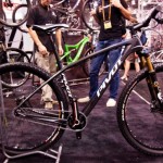A high-performance mountain bike displayed at a bicycle expo, featuring a sleek black frame with the brand name "Pivot" prominently displayed. The bike is on a stand, showcasing its front suspension and intricate design details. In the background, attendees are interacting and other bikes can be seen. The setting is a crowded exhibition hall with a focus on cycling technology.