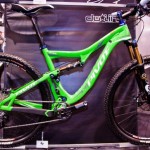 Image of a bright green mountain bike with a sturdy frame and modern design. The bike features thick tires, a prominent front suspension fork, and branded components. It is displayed against a dark background at an exhibition, highlighting its vibrant color and sleek lines.