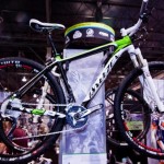 A sleek mountain bike on display at a trade show, featuring a black and green frame, large black wheels with red and white accents, and a modern design. The backdrop includes promotional banners and other cycling equipment.