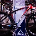A red and black mountain bike displayed on a stand at an exhibition, featuring a sleek design and the brand name "Fuji" prominently on the frame and tires. The background includes colorful banners and blurred figures of attendees, indicating a vibrant event atmosphere.