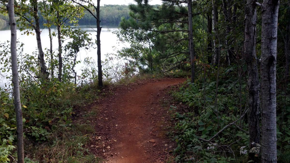 A winding dirt path surrounded by trees leads towards a calm lake, creating a serene natural landscape. The scene features greenery and foliage, with the water reflecting the surrounding forest. Cuyuna Lakes mountain bike trail.