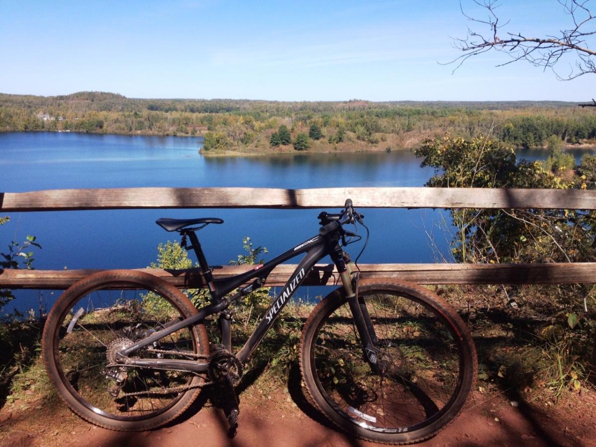 A mountain bike rests against a wooden railing overlooking a serene blue lake surrounded by lush greenery and hills under a clear blue sky. Cuyuna Lakes mountain bike trail.