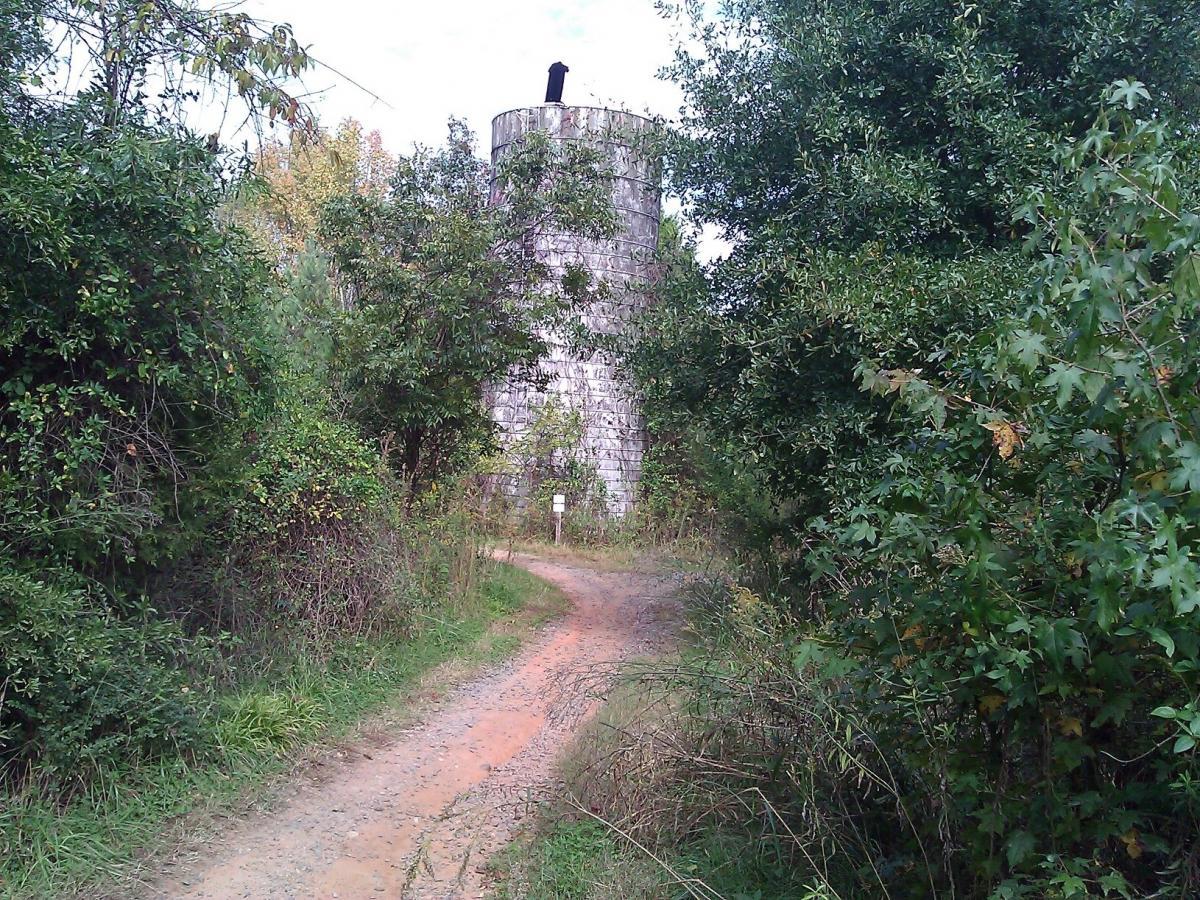A narrow dirt path winding through lush greenery, leading to an old, round stone silo surrounded by trees and shrubs. The sky is partly cloudy, and the top of the silo is visible against the foliage. Sherman Branch Park Mtb Trail mountain bike trail.