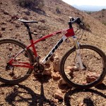 A red and white mountain bike positioned on rocky terrain, with desert vegetation and rolling hills visible in the background under bright sunlight. The bike features thick tires suited for off-road riding and a sturdy frame.