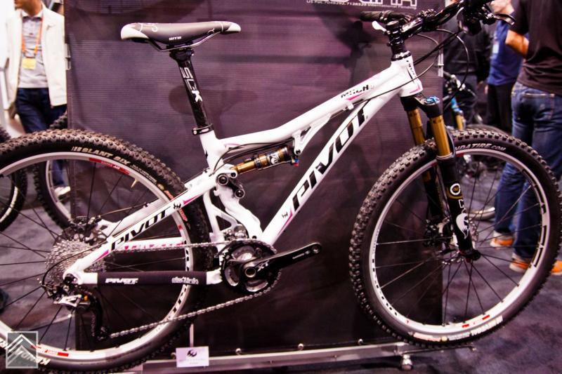 Pivot Mach 4: A white Pivot mountain bike displayed upright on a stand, featuring a sleek frame, wide tires, and front suspension. The bike showcases components like a chainring and a suspension system, with a crowd of people in the background, highlighting its presence at a biking event.