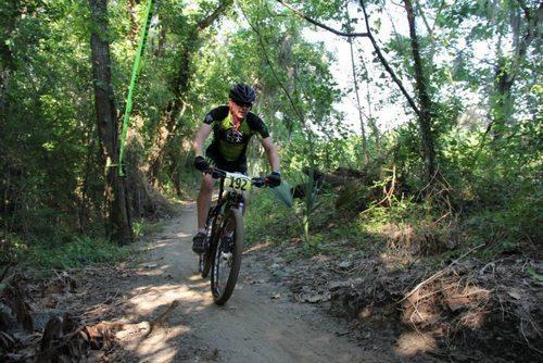 A person riding a mountain bike on a dirt trail surrounded by lush green trees. The cyclist is wearing a helmet and biking gear, with a race number visible on their shirt. Sunlight filters through the foliage, highlighting the natural setting. Bonnet Carre Spillway Trail mountain bike trail.