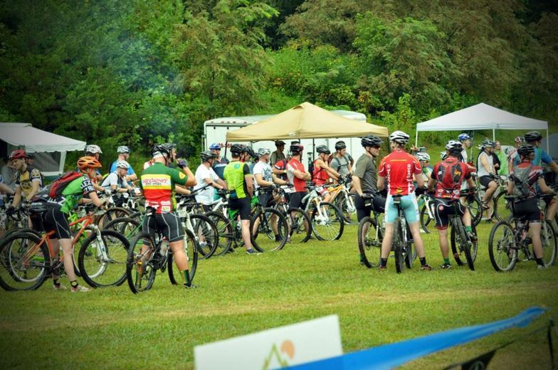 A group of cyclists gathered in a grassy area, with many wearing helmets and colorful cycling jerseys. They are standing beside their mountain bikes, conversing in anticipation of an event. Tents are set up in the background, surrounded by trees and greenery. Woolwine Trails [Shiners Revenge] mountain bike trail.