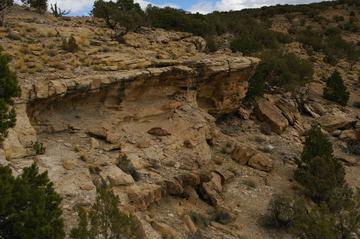 A rocky landscape featuring a prominent cliff edge with a flat overhang, surrounded by sparse vegetation and rugged terrain under a cloudy sky. Sidewinder mountain bike trail.