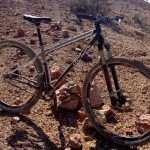 A mountain bike positioned on rocky terrain with a desert landscape in the background. The bike features a silver frame, wide tires, and a black saddle. The ground is covered with stones and sparse vegetation, under clear blue skies.