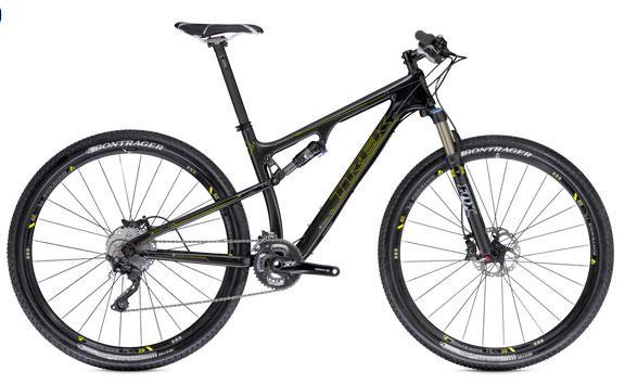 Trek Superfly 100 Elite carbon: A black mountain bike with a lightweight frame and front suspension, featuring disk brakes and knobby tires. The bike has a sleek design suitable for off-road riding, with gear components visible near the pedals.