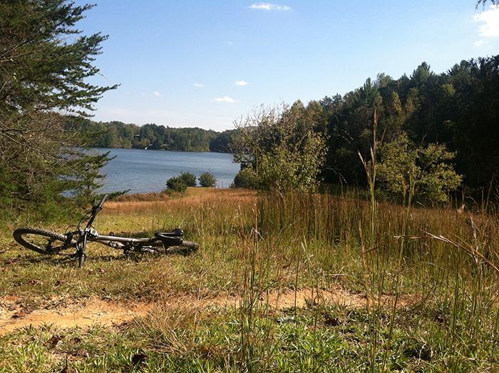 A scenic view of a tranquil lake surrounded by greenery and trees, with a dirt path leading toward the water. A mountain bike is lying in the foreground on a grassy area, framed by tall grass and autumn foliage under a clear blue sky. Warrior Creek mountain bike trail.