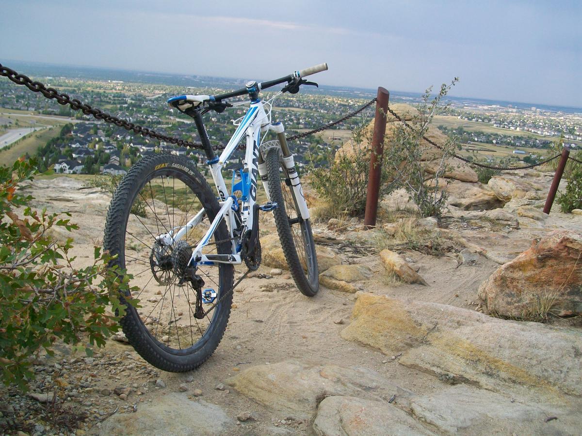 Giant Anthem X 29er 3: A mountain bike parked on rocky ground overlooking a scenic view of a town and surrounding landscape, with a chainlink fence in the background and clouds in the sky.