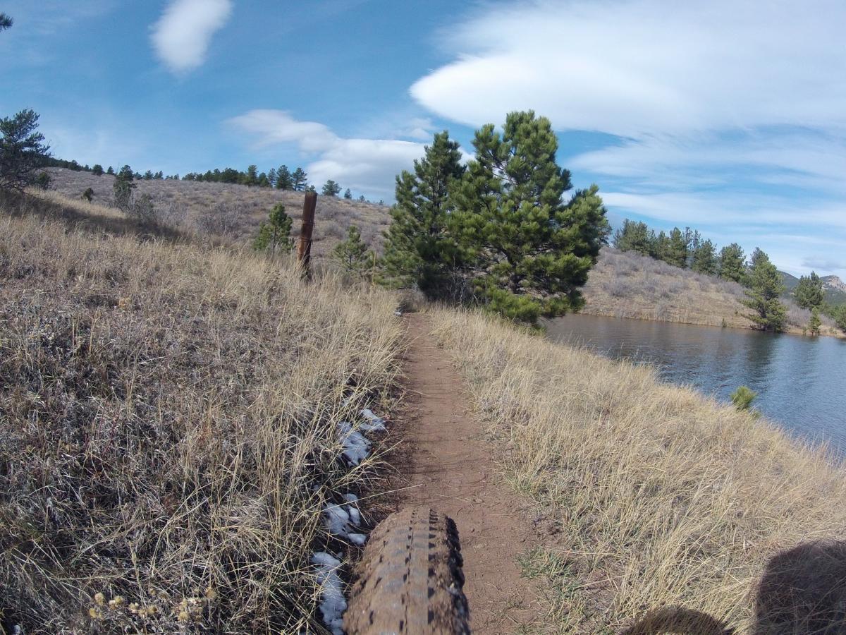 A dirt path runs alongside a calm lake, bordered by sparse grass and a few scattered pine trees. In the foreground, the tire of a mountain bike is visible, suggesting an outdoor adventure. The sky features white, wispy clouds, and the landscape is hilly in the background. Pinewood Reservoir mountain bike trail.