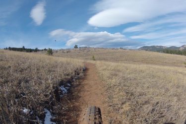 A dirt trail winding through a grassy landscape with low vegetation, under a blue sky with white clouds. The view includes a distant hill and mountains, suggesting a natural outdoor setting. Pinewood Reservoir mountain bike trail.