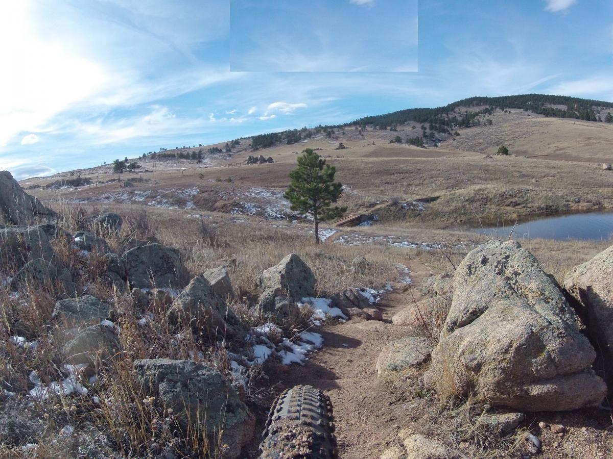 A scenic landscape featuring rocky terrain, grassy fields, and a small pond. In the foreground, there's a bike tire track leading the way, with large rocks and sparse vegetation visible. The background consists of rolling hills dotted with trees under a clear blue sky with scattered clouds. The area shows signs of winter with patches of snow on the ground. Pinewood Reservoir mountain bike trail.