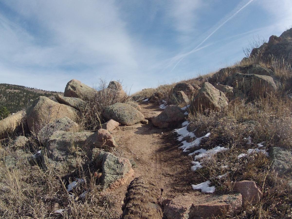 A dirt trail winding through rocky terrain, lined with boulders and sparse grass. In the background, a panoramic view of rolling hills and a blue sky with wispy clouds can be seen. Some patches of snow are visible along the trail. Pinewood Reservoir mountain bike trail.