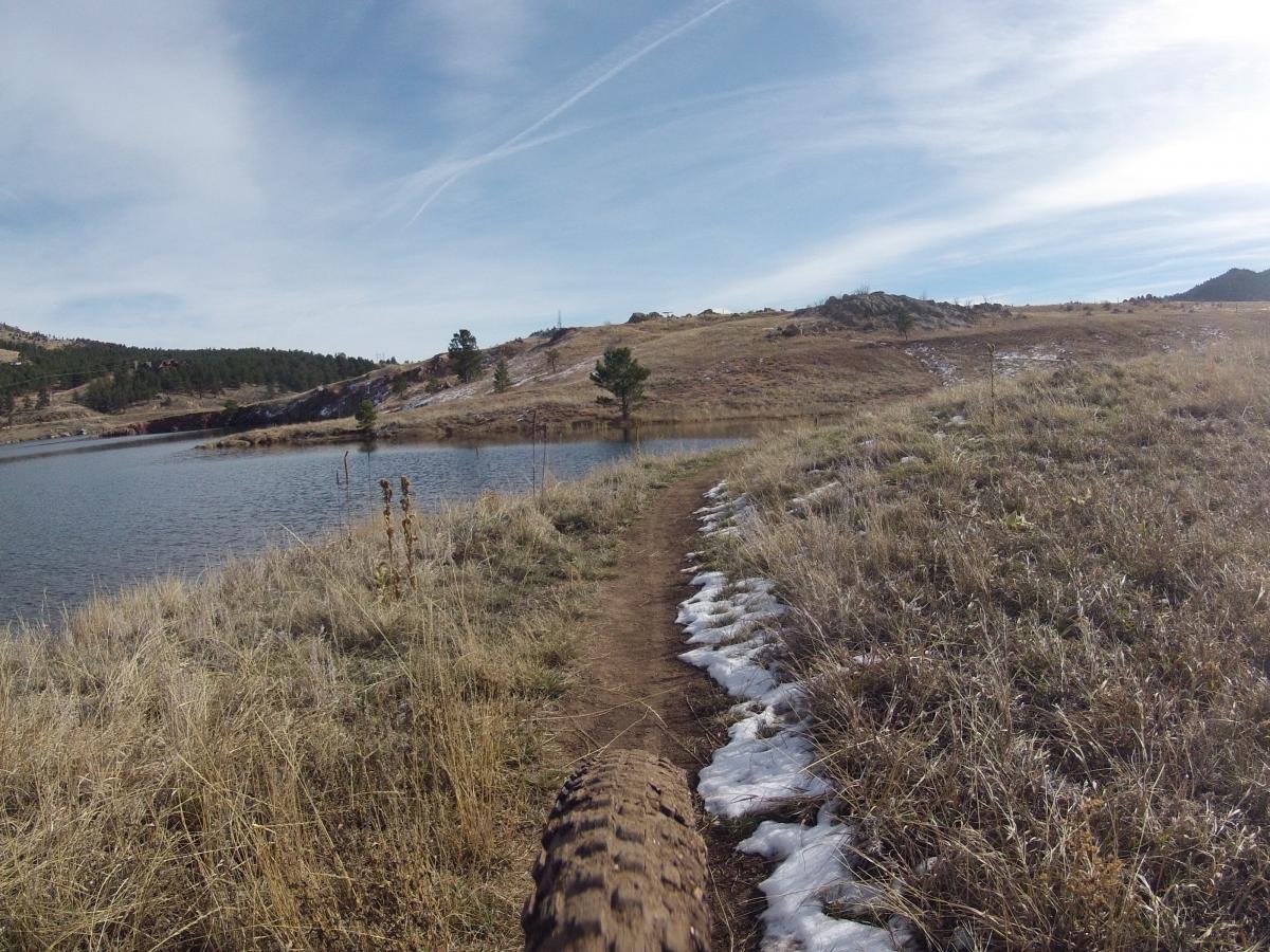 A scenic view of a dirt trail winding along the edge of a lake, surrounded by grassy hills and scattered pine trees. The sky is clear with wispy clouds, and patches of snow are visible on the ground, indicating a cool climate. Pinewood Reservoir mountain bike trail.