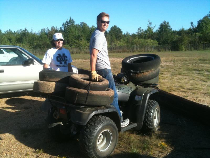 A man and a woman are standing next to an ATV loaded with several used tires. The man, wearing a gray t-shirt and sunglasses, smiles at the camera while seated on the ATV. The woman, dressed in a white hat and a t-shirt, stands beside the ATV, also smiling. In the background, there are trees and a parked car. The scene is set in an open area, reflecting a sunny day. Butts Park mountain bike trail.