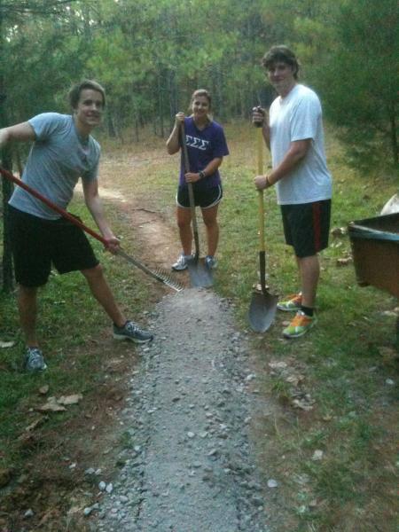 Three young adults are standing in a wooded area, each holding gardening tools. Two of them are holding shovels, while one has a rake. They are working on a gravel path that is being constructed. The scene conveys a sense of teamwork and outdoor activity, with trees and greenery in the background. Butts Park mountain bike trail.