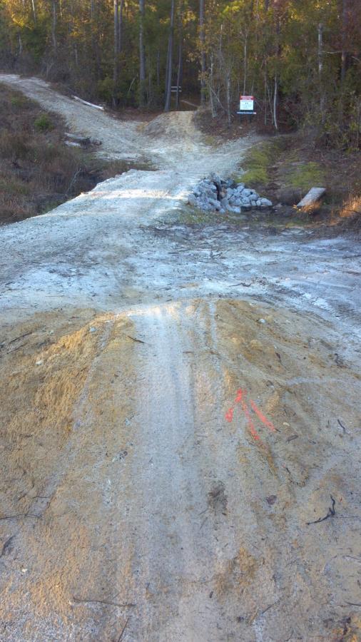 A dirt path leading down to a rocky stream bed, with signs of erosion and sand. In the background, there are trees and a warning sign visible on the right side of the path. Brunswick Nature Park mountain bike trail.