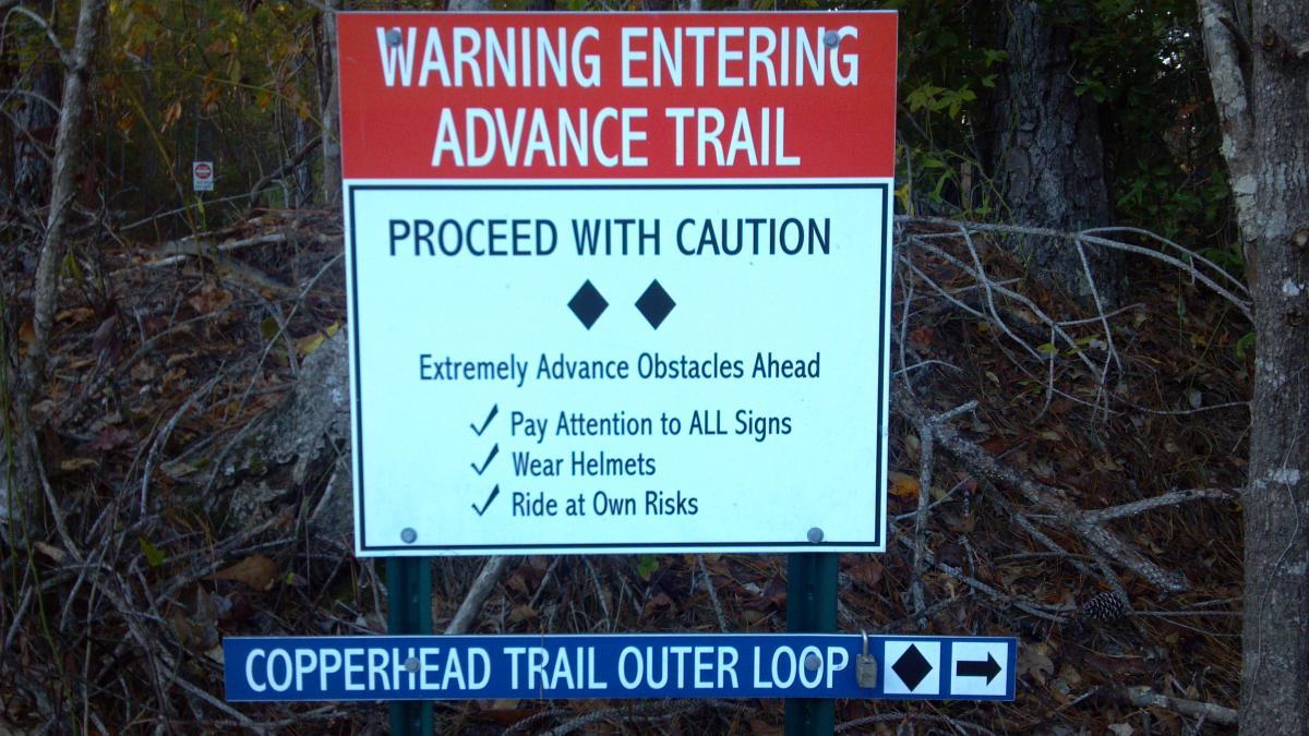 A warning sign indicating entry into an advanced trail. It advises caution and mentions extremely advanced obstacles ahead, along with guidelines: pay attention to all signs, wear helmets, and ride at own risk. Below the warning, there is a directional sign for the Copperhead Trail Outer Loop. The background features trees and foliage. Brunswick Nature Park mountain bike trail.