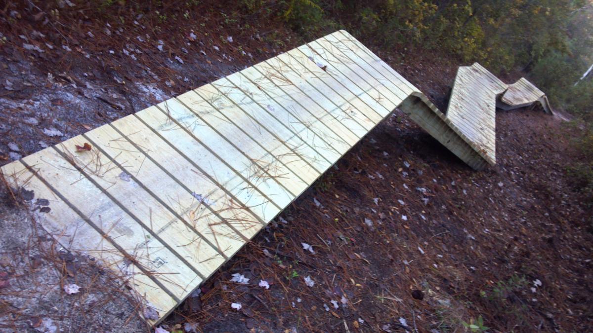 A wooden pathway that appears to be weathered and uneven, surrounded by a forest floor covered in pine needles and scattered leaves. The boards are disjointed and partially raised, suggesting potential wear or damage to the structure. Brunswick Nature Park mountain bike trail.