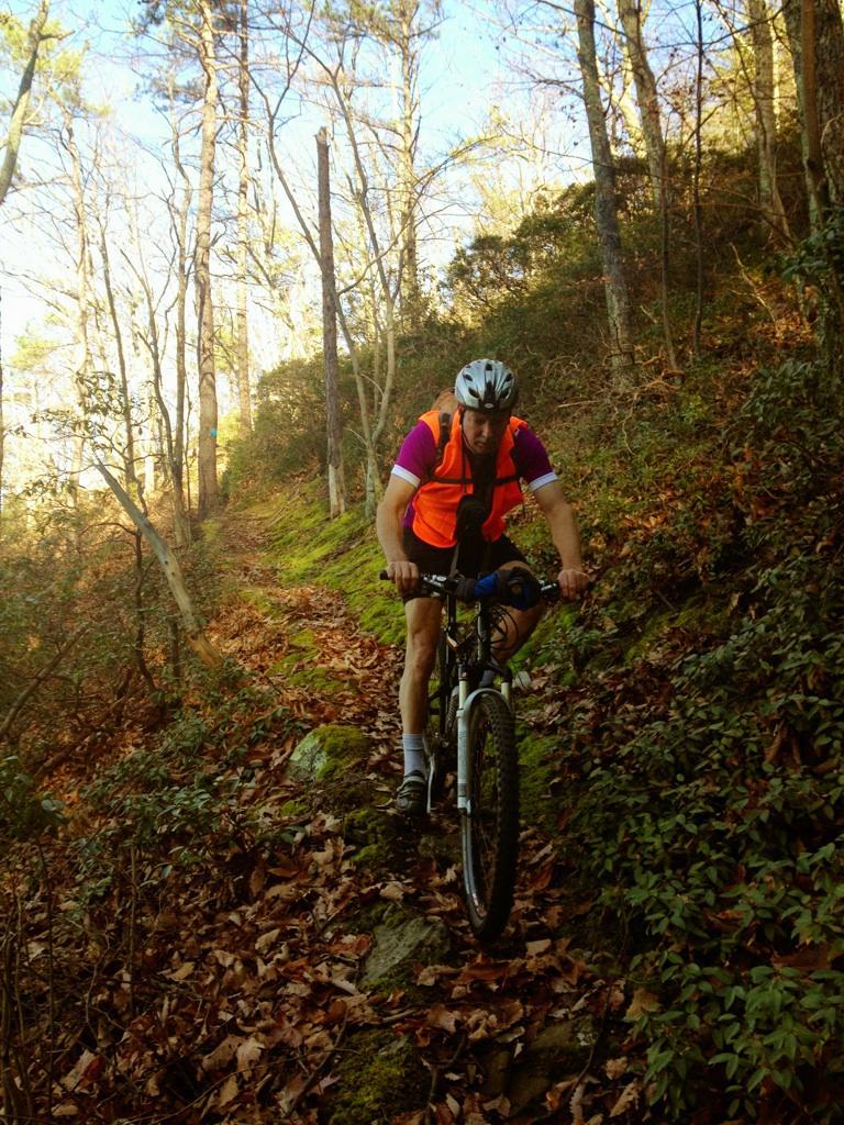 A cyclist wearing an orange vest navigates a rocky trail surrounded by trees and fallen leaves, showcasing an outdoor setting perfect for mountain biking. Dragon's Back mountain bike trail.