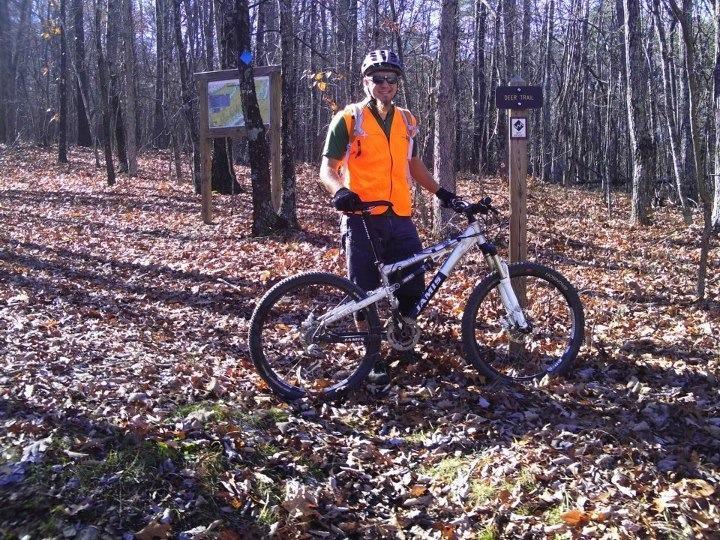 A person wearing an orange safety vest and a helmet stands next to a mountain bike in a wooded area covered with autumn leaves. Behind them, there is a trail sign labeled "Derby Trail" and a map display board, indicating a mountain biking trail in a forested setting. Sunlight filters through the trees, creating a bright and inviting atmosphere. Dragon's Back mountain bike trail.