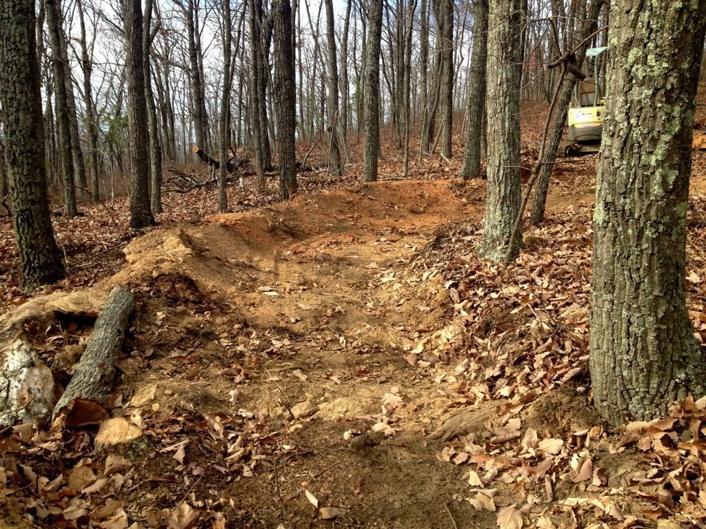 A dirt path through a forested area, featuring exposed soil and fallen leaves. Several trees with bare branches are visible in the background, indicating a seasonal change. A small construction vehicle is partially visible on the right side of the image. Split Decision mountain bike trail.