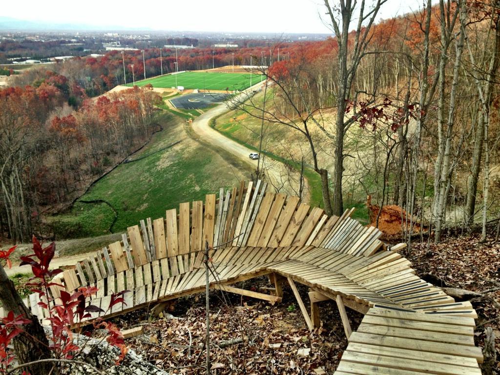 A wooden spiral pathway winding down a hillside, surrounded by trees with autumn foliage. In the background, a green sports field is visible along with a winding dirt road. The scene captures a blend of natural beauty and human-made structure. Liberty Mountain mountain bike trail.