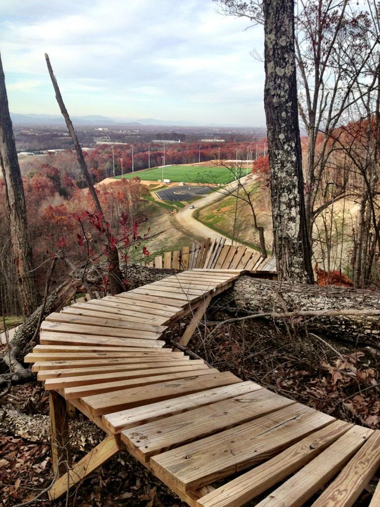 A wooden spiral pathway winding through a forest, overlooking a landscape of rolling hills and a sports field below. The scene is set during autumn, with colorful foliage surrounding the pathway and a cloudy sky above. Liberty Mountain mountain bike trail.