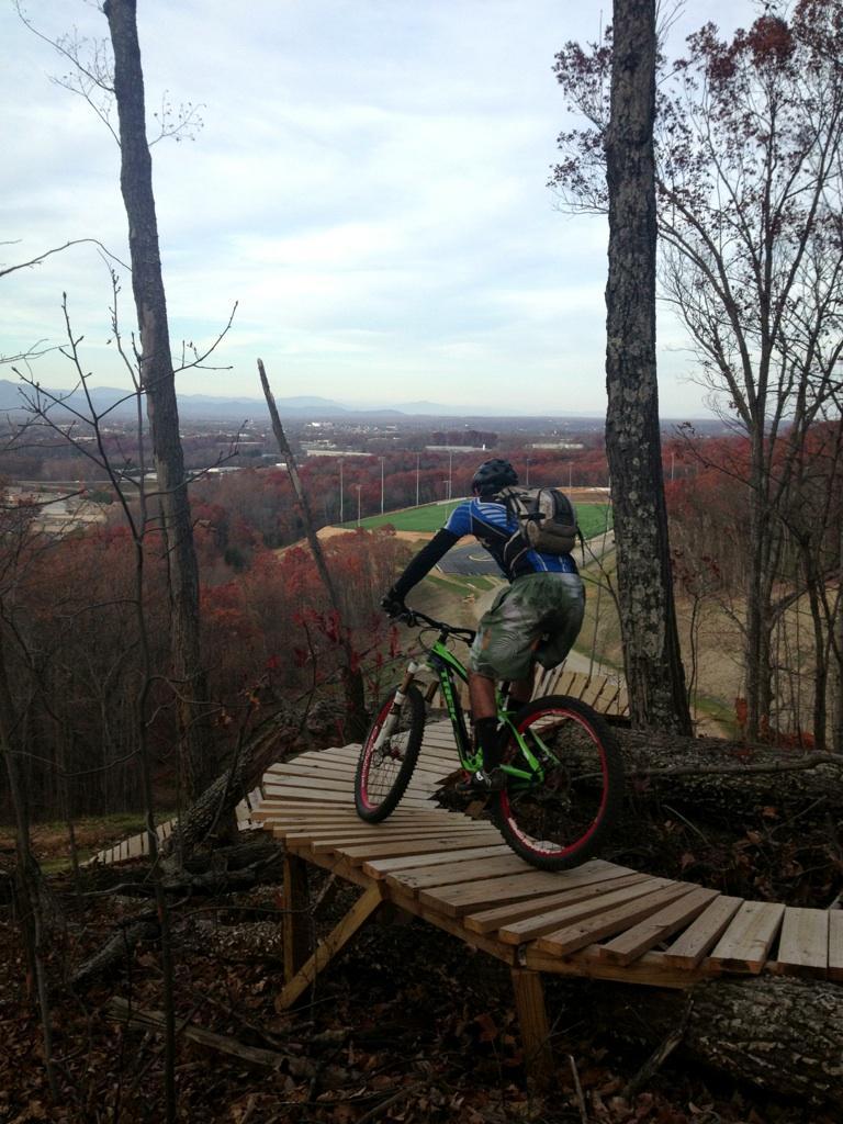 A mountain biker navigating a wooden trail on a hillside, surrounded by autumn foliage. The view in the background shows a valley and fields, with mountains visible in the distance under a cloudy sky. Liberty Mountain mountain bike trail.