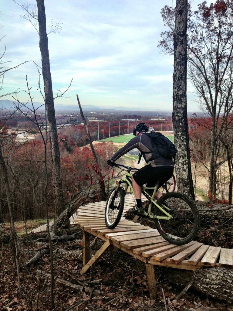 A mountain biker descending a wooden trail bridge through a forest, surrounded by autumn foliage, with a scenic view of the landscape in the background. Liberty Mountain mountain bike trail.