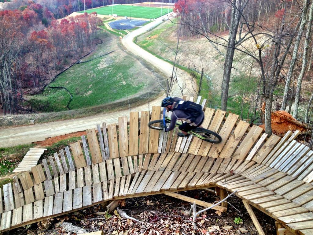 A mountain biker riding on a wooden ramp that curves along a hillside, surrounded by trees with autumn foliage. In the background, a grassy area and a sports field are visible, along with a winding dirt path. Liberty Mountain mountain bike trail.