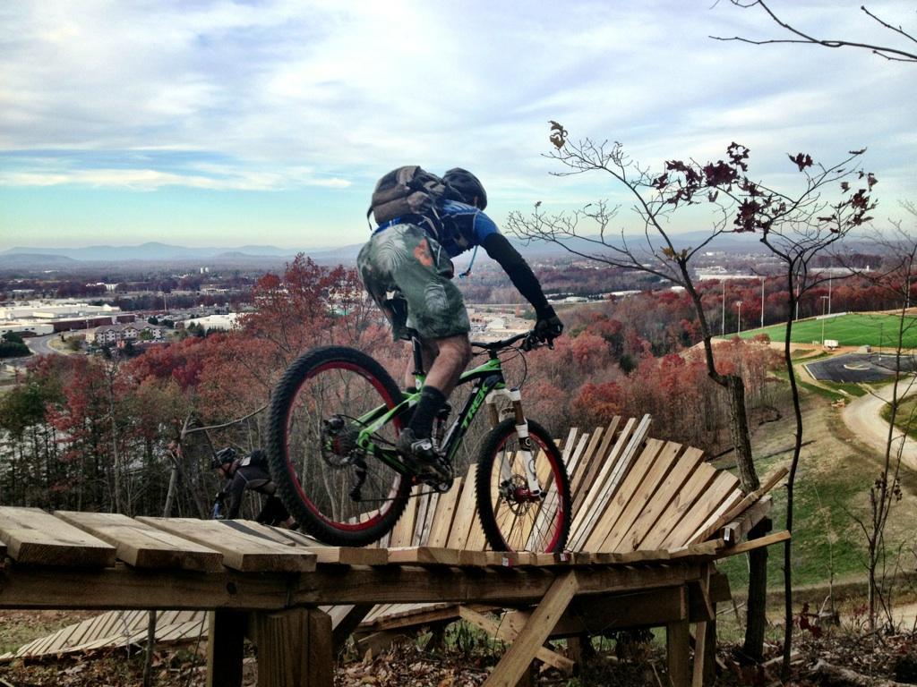 A mountain biker navigating a wooden ramp on a hillside, with scenic views of a valley and autumn foliage in the background. The biker is wearing a backpack and riding a green mountain bike, demonstrating skill as they balance on the structure. Liberty Mountain mountain bike trail.