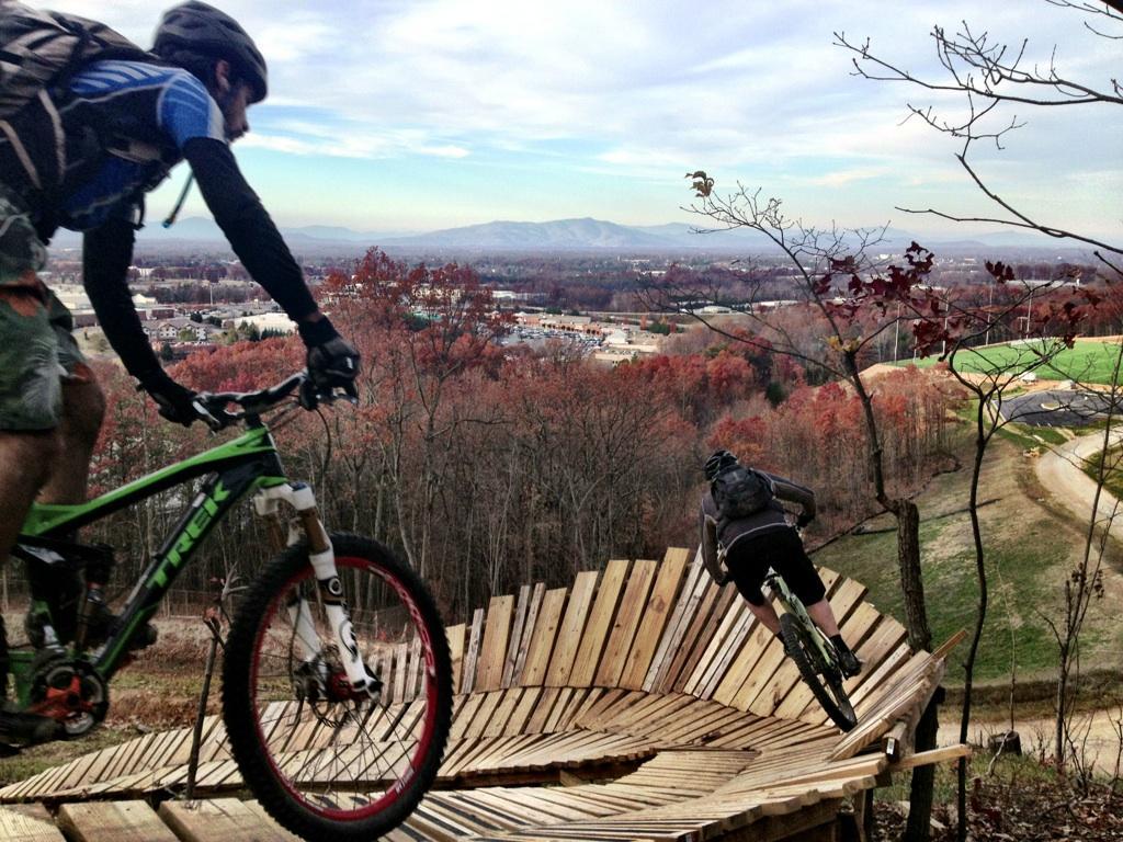 Two mountain bikers navigate a wooden ramp on a hillside, with a scenic view of a valley and mountains in the background. The foliage is in autumn colors, showcasing shades of orange and red, while the sky is partly cloudy. One rider is in a blue shirt and shorts, and the other is in a black shirt and shorts, both wearing helmets. The terrain includes a winding dirt path and a wooden structure designed for biking. Liberty Mountain mountain bike trail.