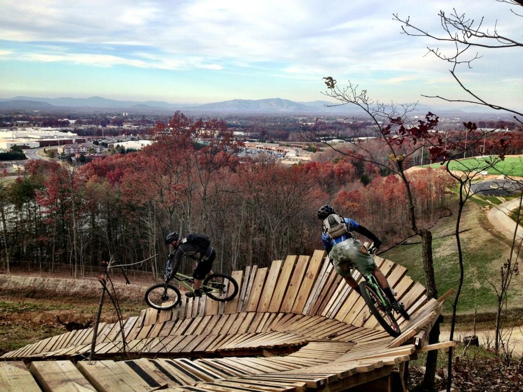 Two mountain bikers navigate a wooden downhill path on a hillside, surrounded by autumn foliage and overlooking a town in the valley below. The sky is cloudy, and the landscape features distant mountains and a mix of urban and natural scenery. Liberty Mountain mountain bike trail.