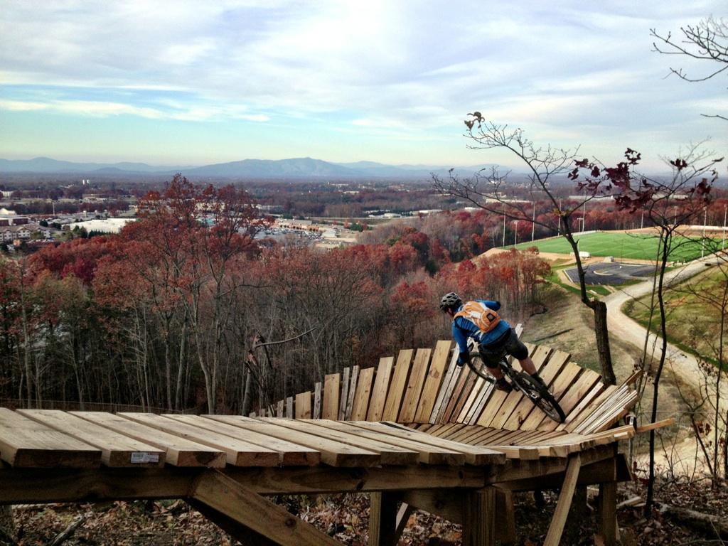 A mountain biker navigates a steep, wooden ramp on a trail, with colorful autumn foliage and distant mountains visible in the background. The scene captures the thrill of outdoor biking in a scenic landscape. Liberty Mountain mountain bike trail.