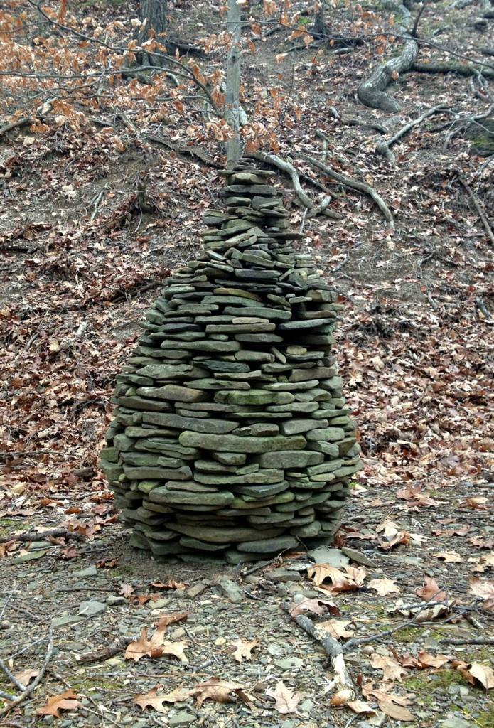 A tall, carefully stacked stone formation resembling a conical shape, set against a backdrop of earthy tones and fallen leaves in a forested area. The structure is made of various sizes of flat rocks, with a tree branch extending from the top. The surrounding terrain shows dry leaves, exposed roots, and a rocky ground. Carvin's Cove Trail system mountain bike trail.