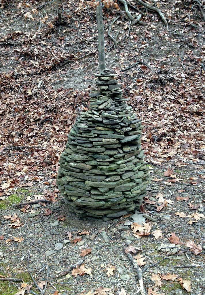A stacked stone sculpture resembling a teardrop shape, created from flat stones and topped with a vertical stick, set against a background of dry leaves and earthy tones. Carvin's Cove Trail system mountain bike trail.