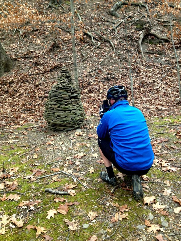 A person in a blue jacket and helmet kneels on the forest floor, photographing a stone cairn made of piled rocks. The background features a wooded area with fallen leaves and bare tree branches. Carvin's Cove Trail system mountain bike trail.