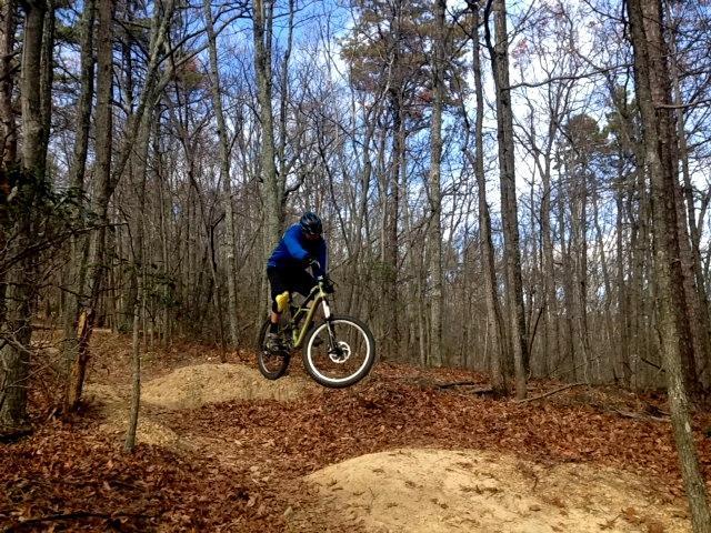 A mountain biker in a blue jacket and helmet is airborne over a dirt jump, surrounded by trees with autumn leaves on the ground. The sky is partly cloudy, showcasing a clear day for biking. Carvin's Cove Trail system mountain bike trail.