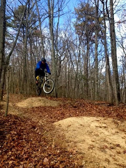 A mountain biker in a blue jacket and helmet executes a jump over a dirt mound on a forest trail, surrounded by trees and fallen leaves. The sky is clear with a few clouds. Carvin's Cove Trail system mountain bike trail.