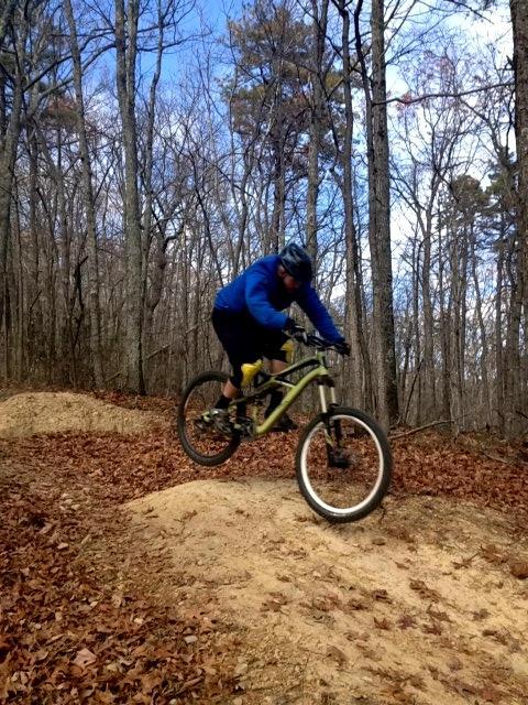 A mountain biker wearing a blue jacket and helmet performs a jump over a dirt mound on a trail surrounded by trees, with autumn leaves on the ground. The sky is partly cloudy, suggesting a clear day for outdoor riding. Carvin's Cove Trail system mountain bike trail.
